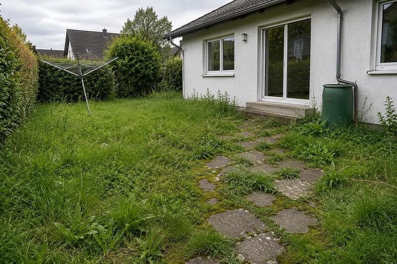 Ungepflegte Gartenfläche mit hohem Gras, alten Betonplatten und einer Wäschespinne